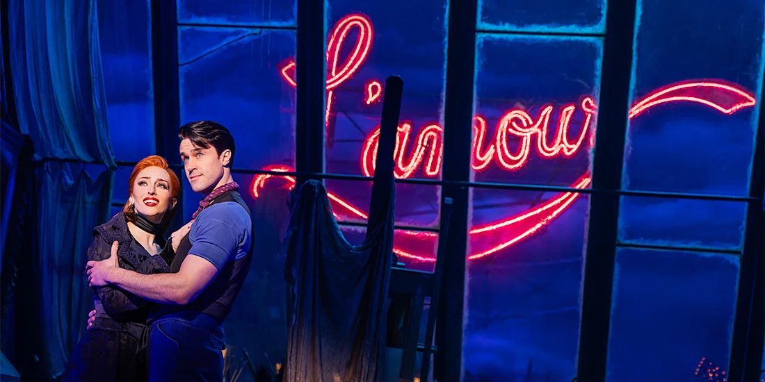 Two actors stand embracing on stage in front of large windows with a glowing red neon "L'amour" sign in the background.
