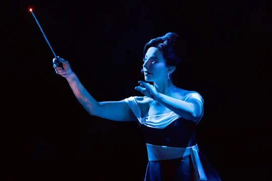 A conductor in an evening gown holding a glowing baton against a dark background.