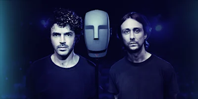 Two men in black shirts stand on either side of a gray, featureless mannequin head with microphones, against a dark, out-of-focus background.
