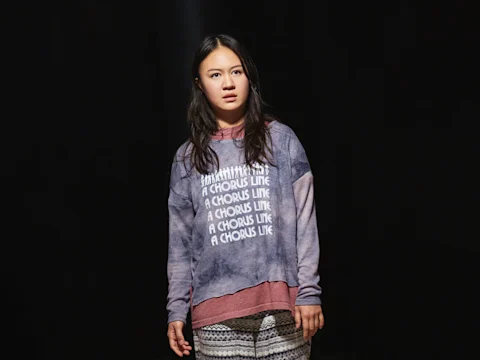 A young woman stands against a dark background, wearing a purple tie-dye sweatshirt that reads "A CHORUS LINE" multiple times and patterned pants, looking slightly to the side.