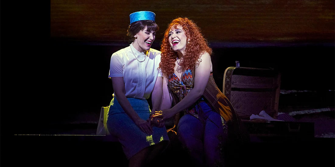 Two women in costume sit together on stage, smiling and holding hands under a spotlight during a theatrical performance.