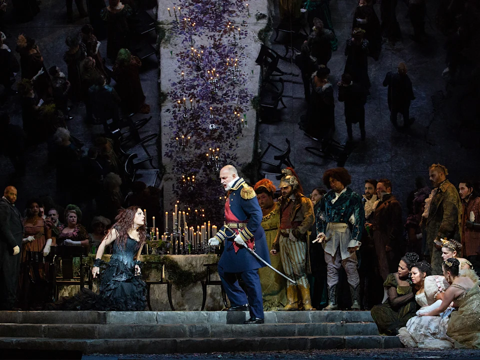 A dramatic opera scene shows a woman kneeling before a uniformed man, surrounded by costumed people, candles, and a long table with flowers.