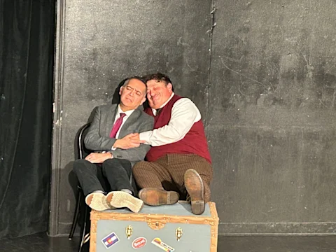 Two men sit closely together on stage; one in a suit, the other in a vest and plaid pants, both leaning on each other with eyes closed, seated atop a trunk decorated with stickers.