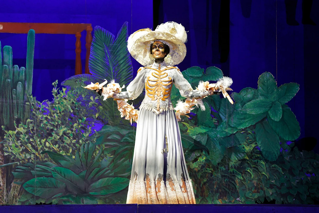 A performer in a white dress and large hat, painted as a skeleton, stands on stage with a jungle-themed backdrop.