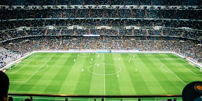 A large soccer stadium filled with spectators shows two teams playing a match on a green field, viewed from a high vantage point.