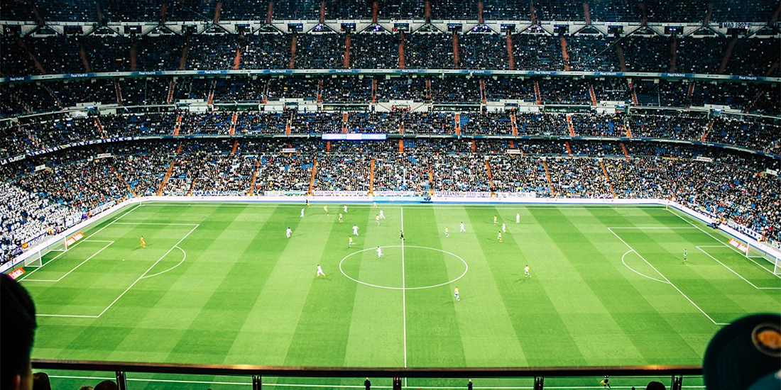 A large soccer stadium filled with spectators shows two teams playing a match on a green field, viewed from a high vantage point.