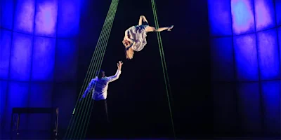 A person in white is suspended upside down midair above a stage, while another person on the ground reaches toward them; blue lights and green laser lines frame the scene.