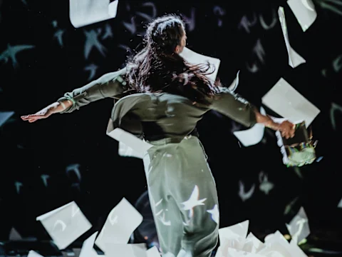 A person in a green outfit twirls amidst flying sheets of paper on a dark background.