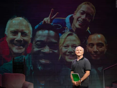 A man holding a notebook stands in front of a wall displaying a projected group photo of five smiling people.