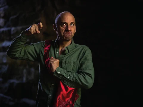 A person in a green shirt poses with a comical expression, gesturing with one hand and holding a red cloth with the other. The background is dark and shadowy.