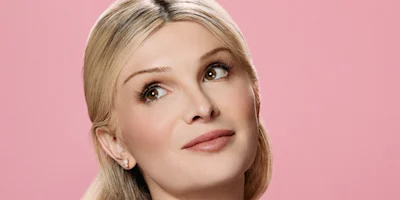 A woman with long blonde hair and light makeup looks slightly upward against a plain pink background.