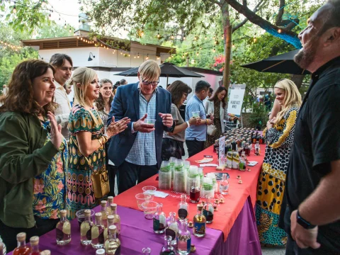 A group of people stands around a table with assorted drinks at an outdoor gathering. Some individuals are engaged in conversation with a person behind the table. Trees and string lights decorate the area.