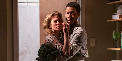 A woman and a man stand closely together in a room, both looking intently ahead with concerned expressions.