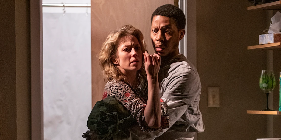 A woman and a man stand closely together in a room, both looking intently ahead with concerned expressions.