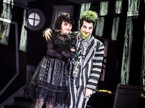 Two people in theatrical costumes stand together on stage; one wears a black dress with lace, the other wears a black-and-white striped suit with green accents and face makeup.