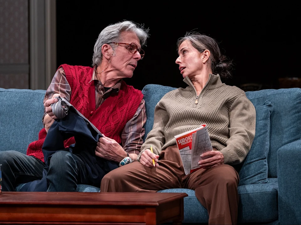 Two adults sit on a blue couch; one holds clothing while the other holds a pen and a Sudoku book, and they appear to be engaged in a serious conversation.