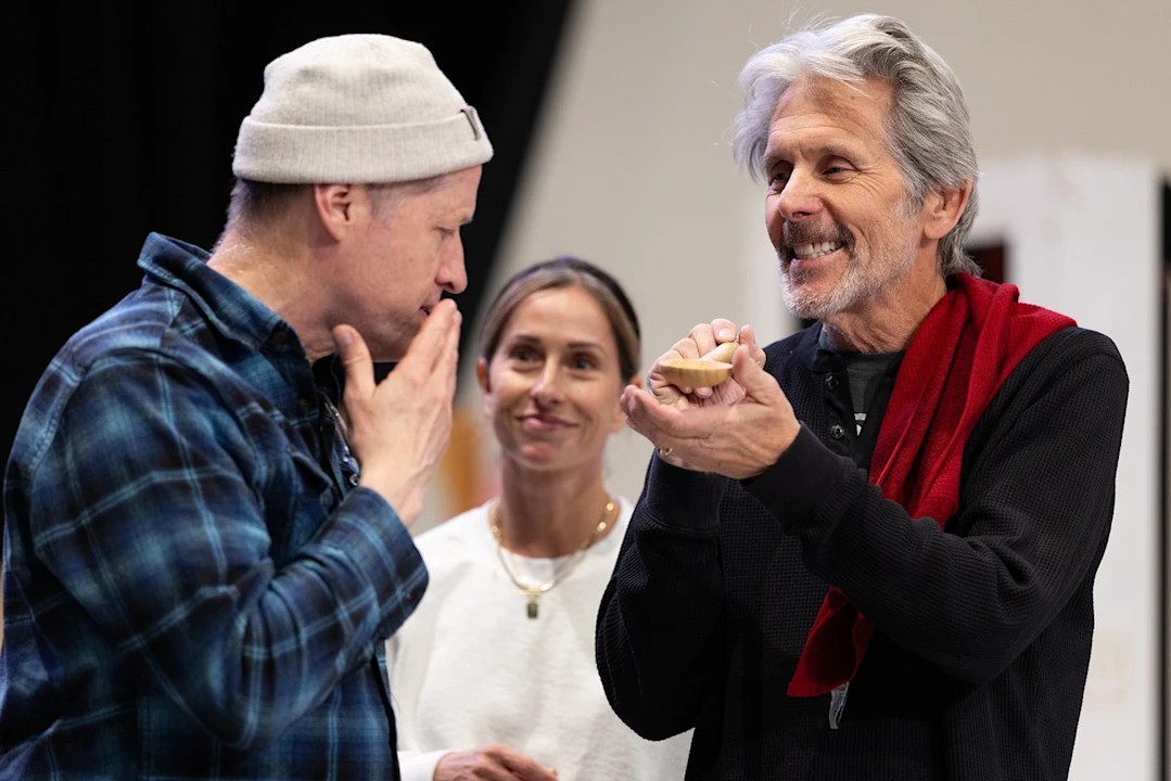 Three people stand together indoors; one man is laughing and holding an object, while another man gestures toward his mouth, and a woman looks on, smiling in the background.