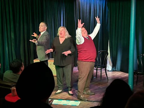 Three actors perform on stage, gesturing expressively, in front of an audience. Black curtains and simple props set the scene for the performance.