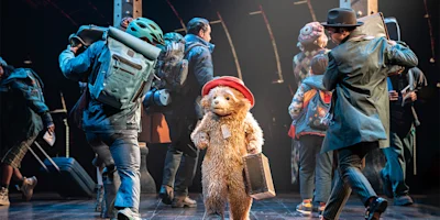 A person in a Paddington Bear costume stands on stage holding a suitcase, surrounded by people in travel attire and backpacks, under spotlights.