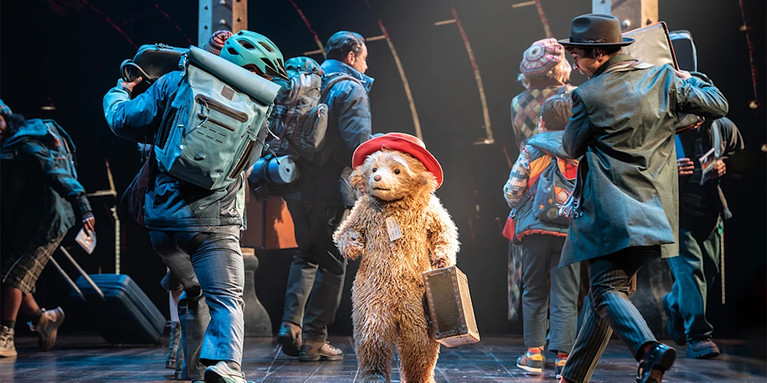 A person in a Paddington Bear costume stands on stage holding a suitcase, surrounded by people in travel attire and backpacks, under spotlights.