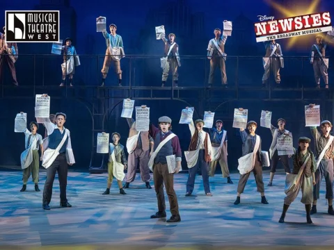 A large group of people dressed as newsboys stand on a stage, holding newspapers in raised hands. Signs for "Musical Theatre West" and "Disney Newsies The Broadway Musical" are visible.