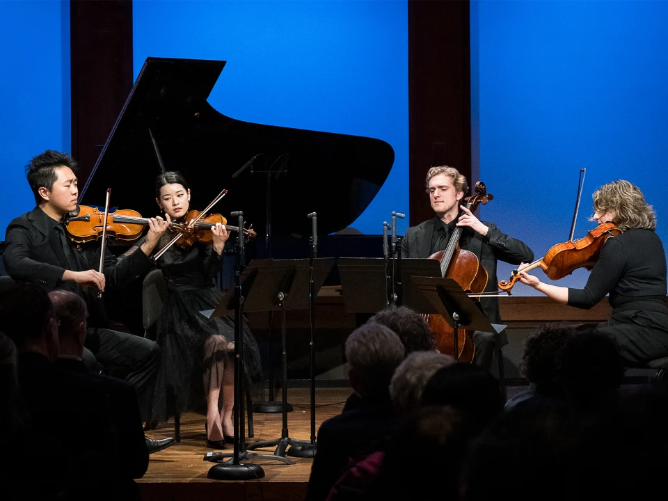 A string quartet and a pianist perform on stage against a blue backdrop. The musicians play the violin, cello, and piano, with sheet music stands in front of them. An audience is seated in the foreground.