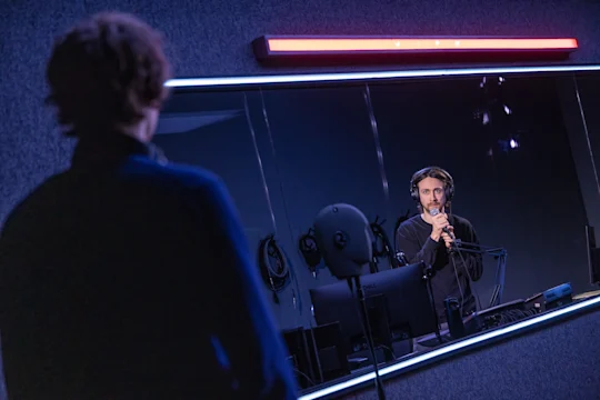 A man in a recording studio wearing headphones and speaking into a microphone, viewed from behind another person.