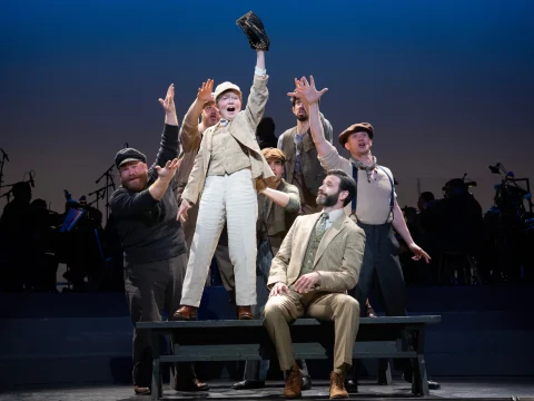 A group of actors perform energetically on stage. One actor stands on a bench, raising a cap, while others reach towards him. An orchestra is visible in the background.