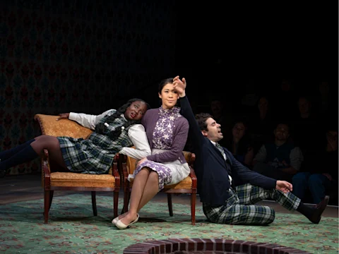 Production photo of The Seat of Our Pants in New York showing, three actors sit dramatically on stage; two wear plaid outfits, one in a purple floral dress, all posed expressively on and around two chairs under stage lighting.