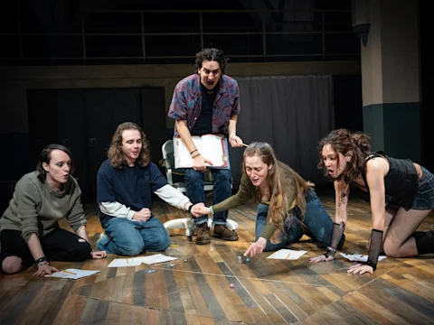 Production photo of Initiative in New York, showing five people are gathered on a wooden floor, intensely focused on dice rolls and papers, with one standing and appearing to narrate or direct the scene.
