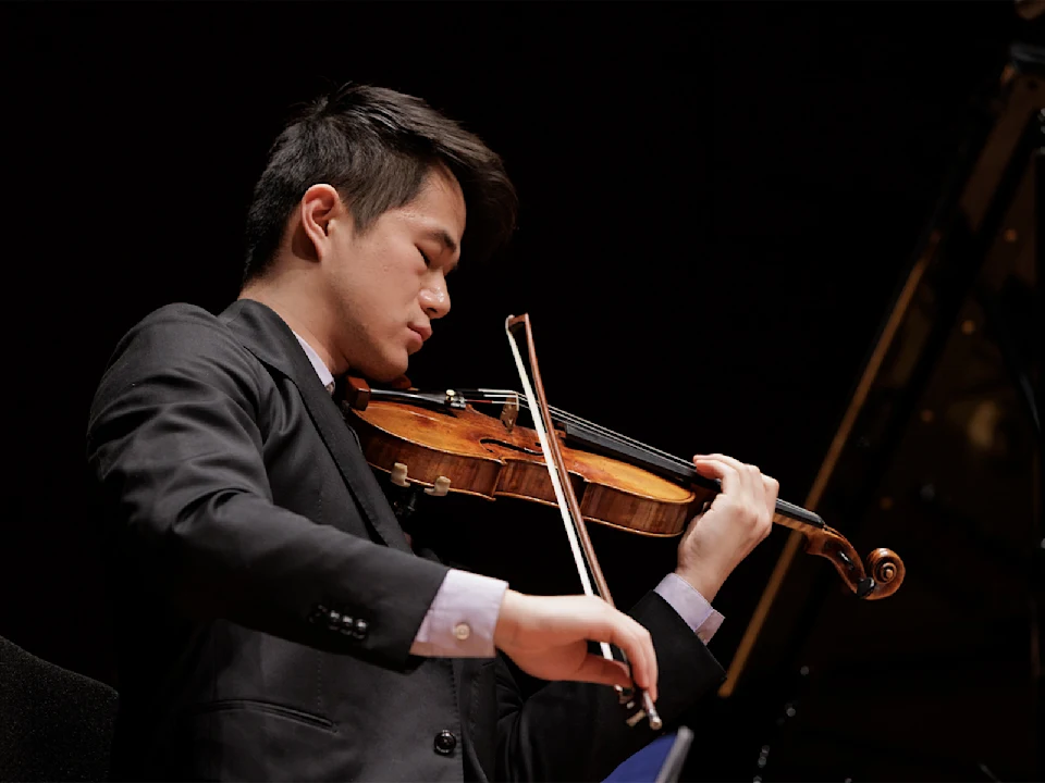 A person in a suit plays a violin on stage, concentrating on the performance, with a dark background.
