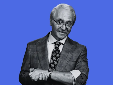 A man with gray hair and glasses wearing a plaid suit jacket and patterned tie stands against a blue background, rolling up his sleeve to reveal a tattoo on his forearm.