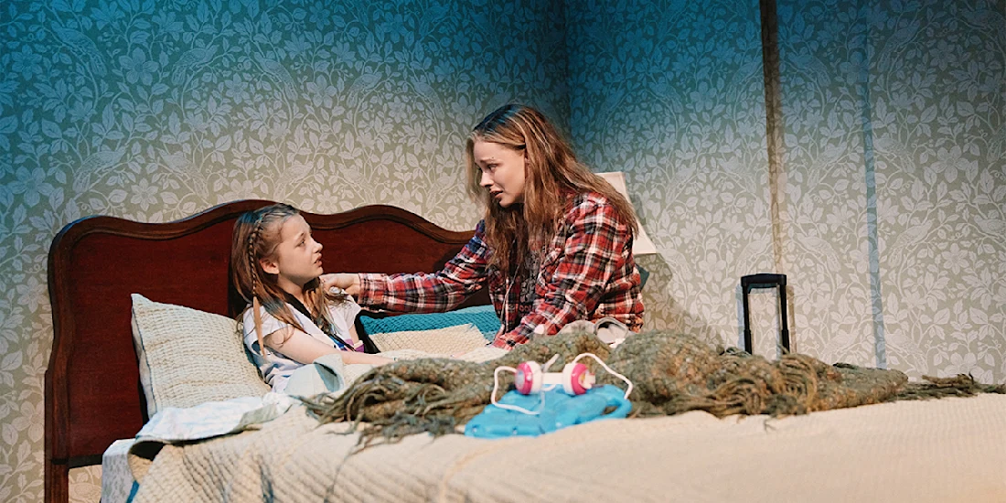 Two girls sit on a bed in a dimly lit room; one, sitting up in bed, listens as the other, wearing a plaid shirt, speaks to her with a concerned expression.