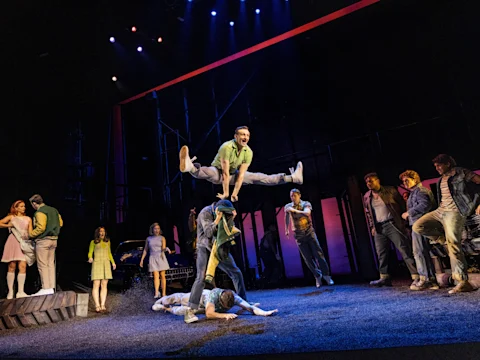 A stage performance with actors in 1960s-style costumes; one actor jumps and splits midair over another actor, while others watch from both sides of the stage.
