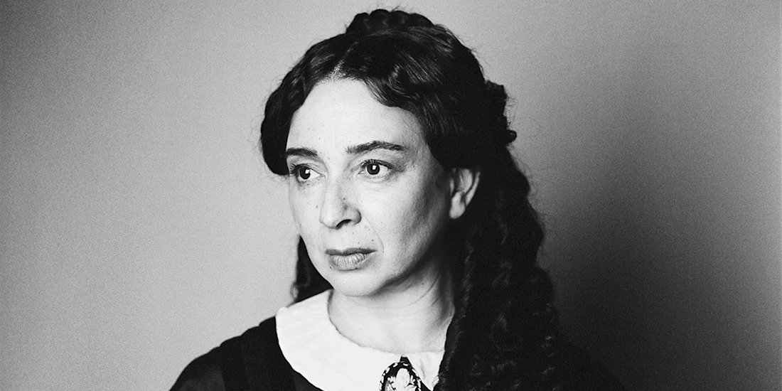 Black-and-white portrait of a woman with long, dark, wavy hair, wearing a white collar and looking slightly to the side with a neutral expression.