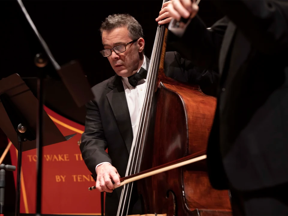 A man in a tuxedo plays a double bass with a bow on stage. Another musician, also in formal attire, is partially visible. Music stands and a red background are seen.