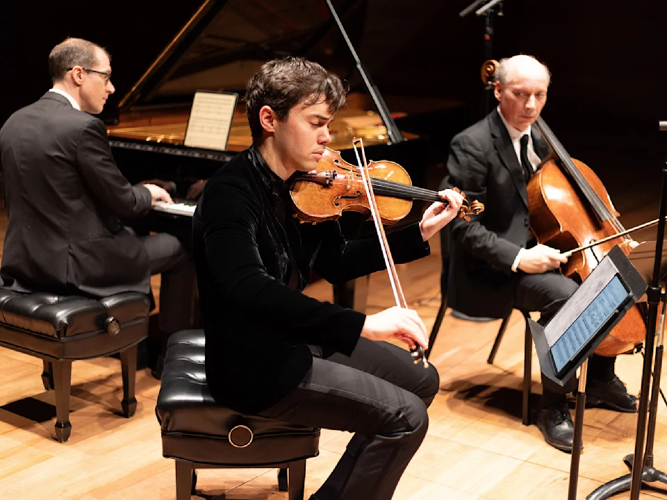 Three musicians perform on stage with a piano, violin, and cello. Each is seated and dressed formally, focused on their instruments and sheet music.