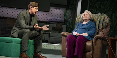 A man in a suit gestures while speaking to an elderly woman seated in an armchair in a domestic setting.