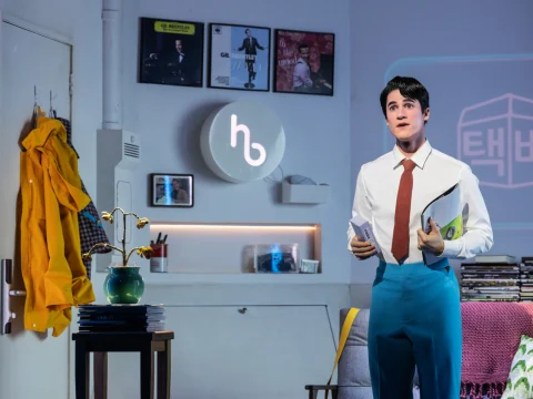 A man in a white shirt and red tie stands in a decorated room holding papers. Behind him are a yellow coat on a hook, wall art, and a round logo with an "h.