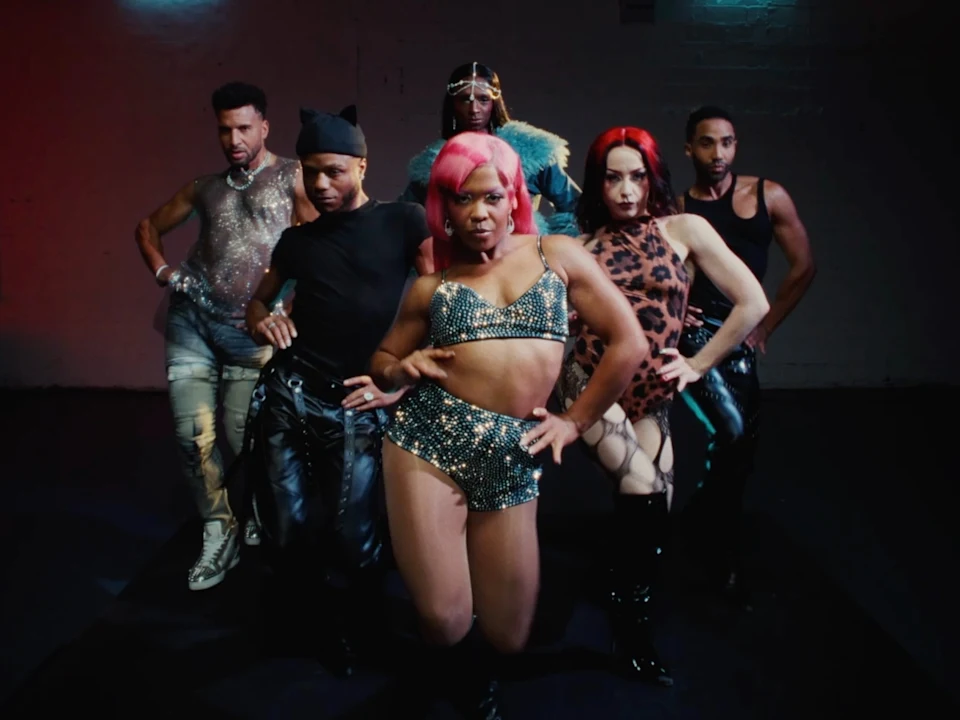 A diverse group of performers in colorful costumes stand on a well-lit stage, some posing and others waving, against a background of vertical blinds.