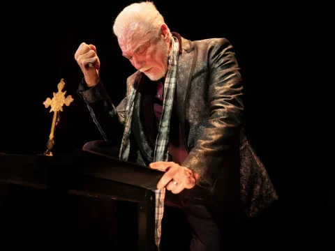 Patrick Page bowing down at a pulpit holding a gold cross, wearing a leather jacket and a plaid scarf.