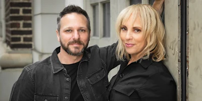A man and woman wearing black shirts stand side by side outdoors, with the woman resting her arm against a wall.