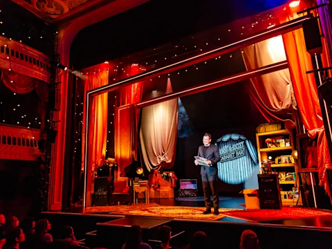 A performer stands center stage in a warmly lit theater set resembling a cozy study, holding books, with an audience watching from the darkened seats.
