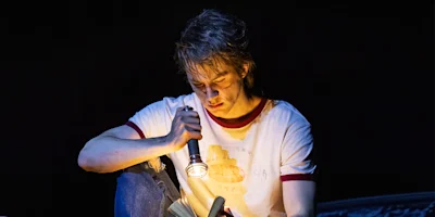A person in a white t-shirt sits in the dark, shining a flashlight on an open book in their lap and reading intently.
