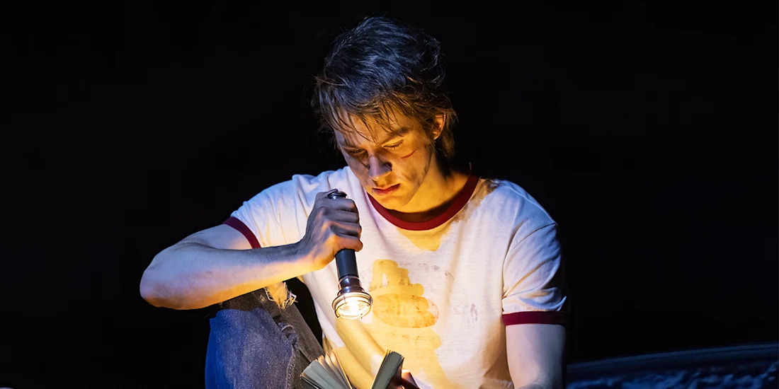 A person in a white t-shirt sits in the dark, shining a flashlight on an open book in their lap and reading intently.