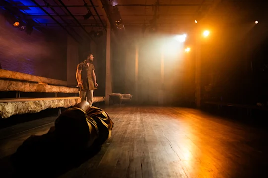 A person stands on a dimly lit stage while another person lies on the floor in the foreground, with warm and cool stage lights illuminating the scene.