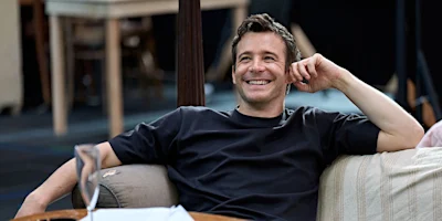 A man in a black t-shirt sitting on a sofa and smiling.