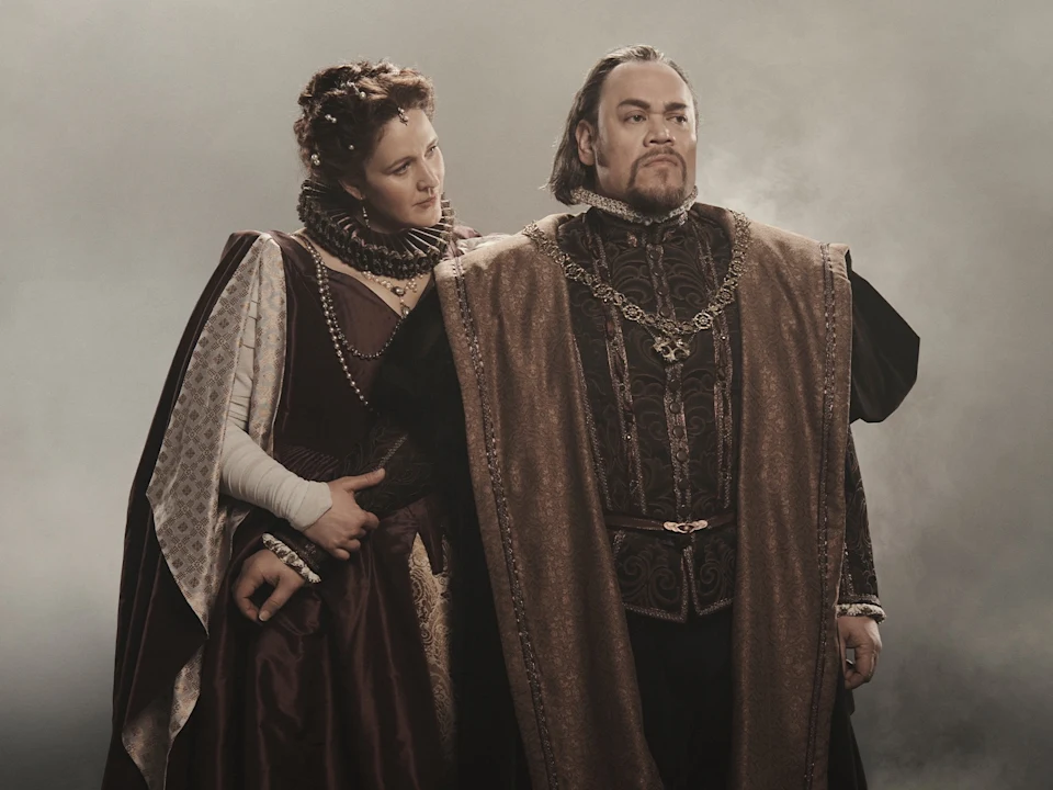 A man and woman in historical royal attire stand together, with the woman looking toward the man.