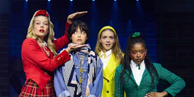 Four young women in colorful, distinct outfits stand on stage under blue lights, with one woman framing another's face with her hands; all have expressive facial reactions.