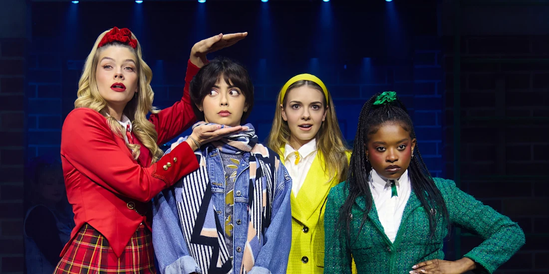 Four young women in colorful, distinct outfits stand on stage under blue lights, with one woman framing another's face with her hands; all have expressive facial reactions.